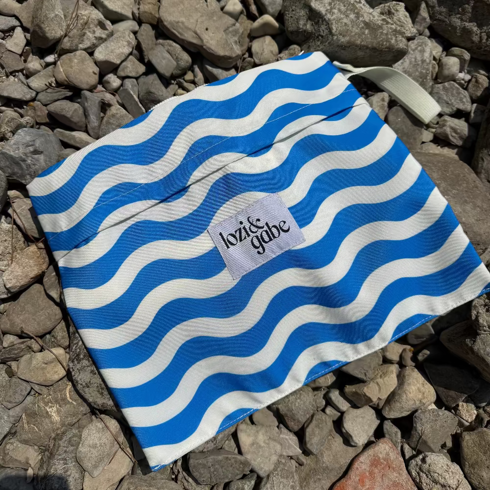 A reusable cloth bag with a blue and white wavy stripe pattern, featuring a small white label that reads 'lozi and gabe' and a white strap, placed on a bed of gray rocks.