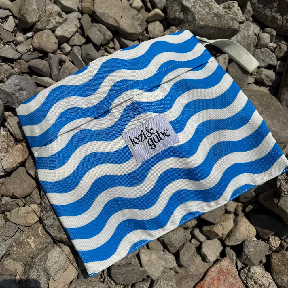 A reusable cloth bag with a blue and white wavy stripe pattern, featuring a small white label that reads 'lozi and gabe' and a white strap, placed on a bed of gray rocks.