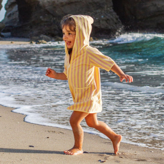 Child in a yellow pocho running on a sandy beach with rocky cliffs and ocean waves in the background.